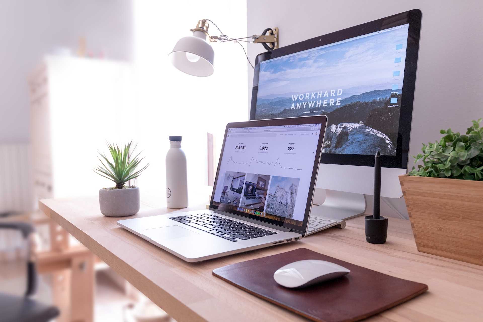 A desk with a desktop computer and a laptop in front of it. There's a white mouse, a white wall lamp, a white water bottle and a couple of plants too. 